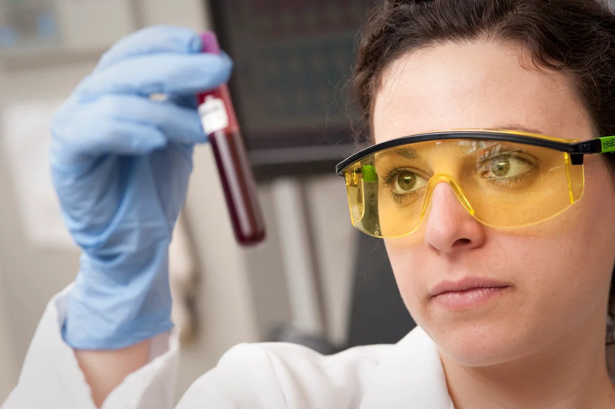 A laboratory scientist wearing gloves and safety glasses examining a tube containing a blood sample.